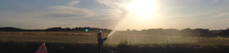 Feuerwehrmann spritzt mit Wasser aus dem Hohlstrahlrohr. Foto mit Gegenlicht der Abendsonne aufgenommen.