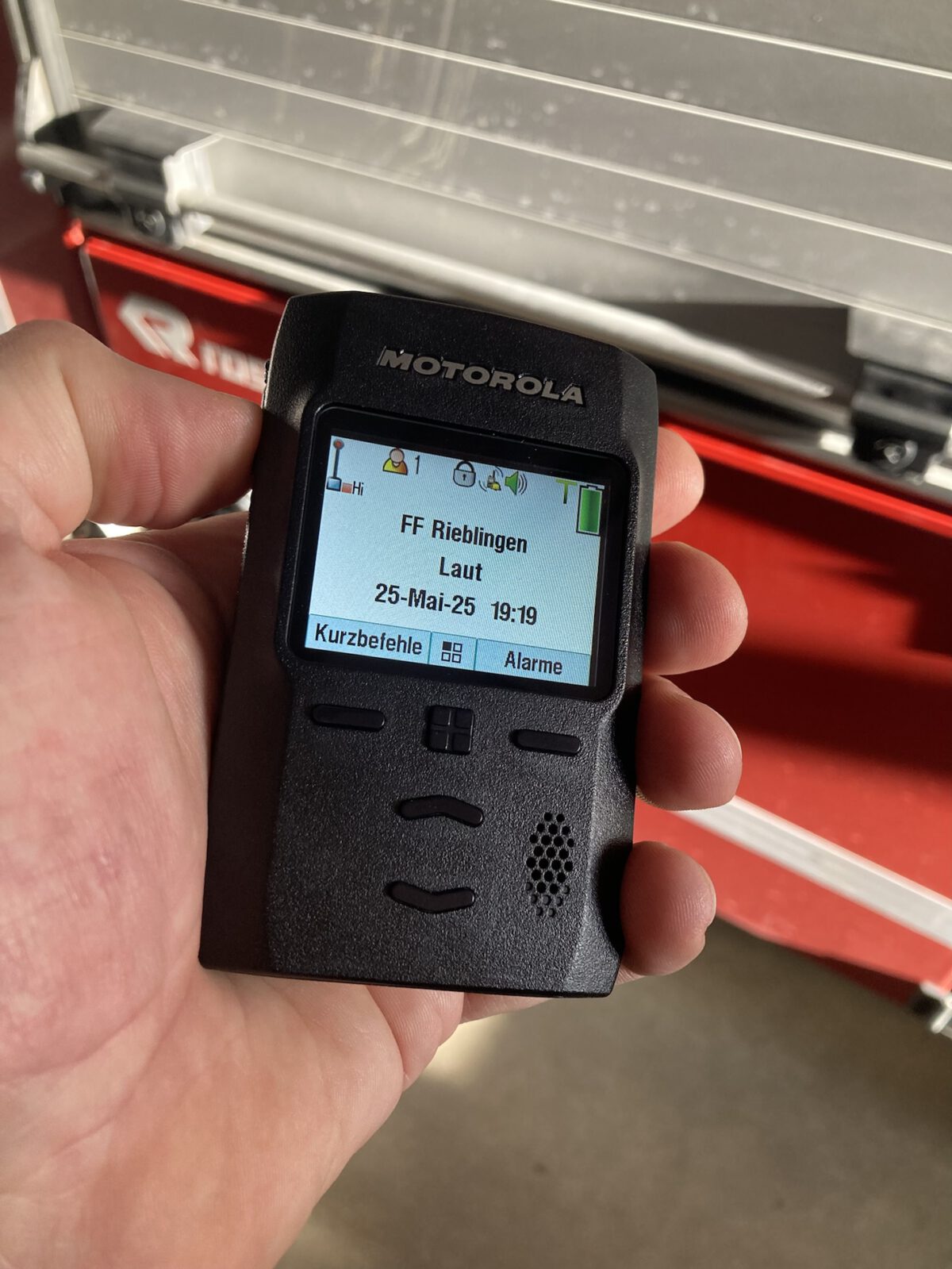 Ein digitaler Funkmeldeempfänger der Firma Motorola. Das Gerät wird in der Hand gehalten, im Hintergrund ist das Feuerwehrauto zu sehen.