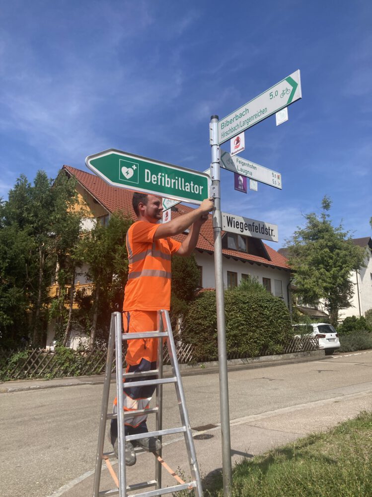 Martin Thoma vom Betriebshof der Stadt Wertingen befestigt das Hinweisschild zum Defibrillator an einer Stange neben der Rieblinger Bushaltestelle.