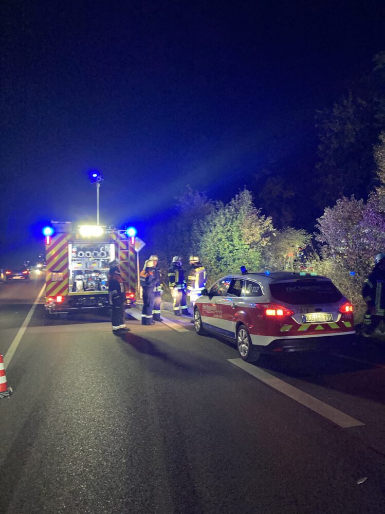 Dunkelheit: Ein hell erleuchtetes Feuerwehrfahrzeug steht auf der Straße, dahinter Feuerwehrleute. Weiter rechts am Straßenrand ein First-Responder-Einsatzfahrzeug Pkw.