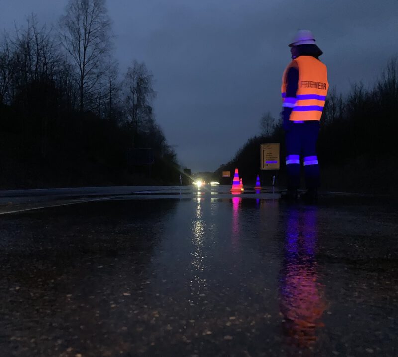 Eine Feuerwehrfrau steht mit oranger Warnweste auf der regennassen Umgehungsstraße und regelt den Verkehr.